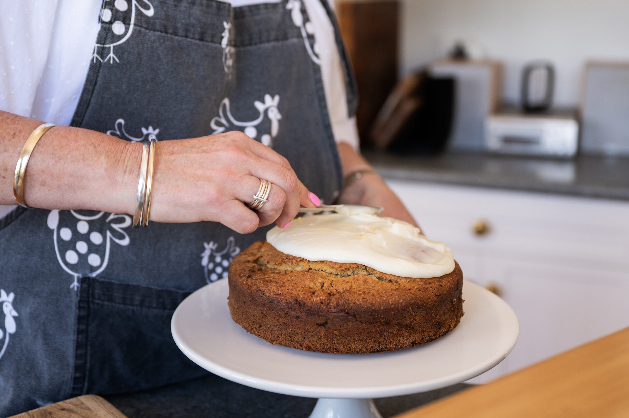 A woman wearing an apron icing a Banana Cake with White Icing
